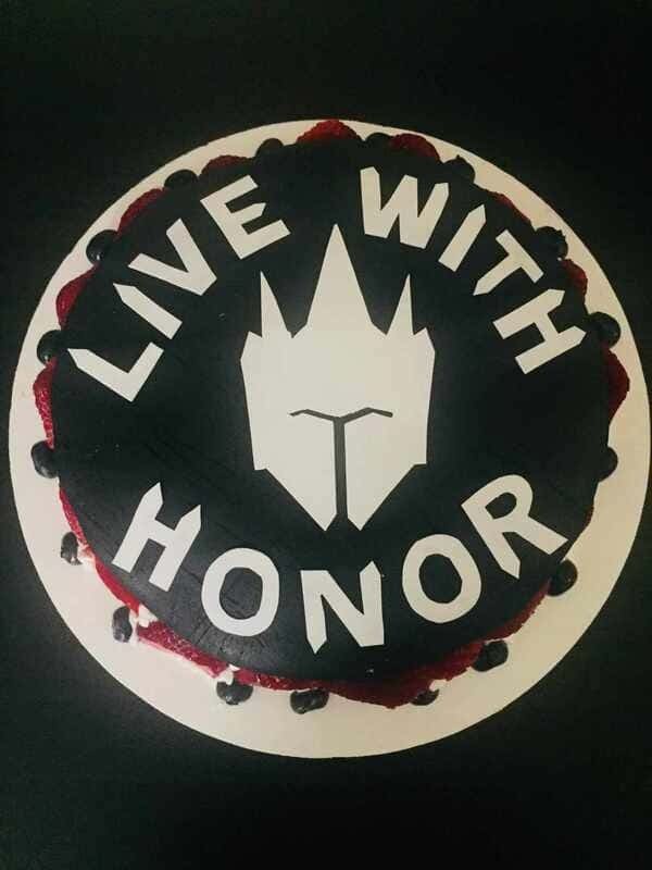 This image shows a cake covered in black fondant and white fondant decorations which write out 'Live with Honor' and a knight's mask.
